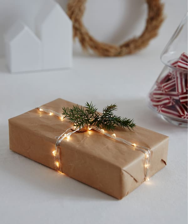 A rectangular gift wrapped in brown paper with a thin, lit lighting chain acting as string; a spruce-like twig for decoration.