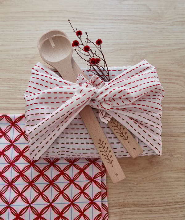 A rectangular gift wrapped not in paper but in a tied piece of cloth, decorated with wooden utensils and a blooming twig.