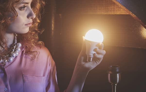 A red-haired woman holding an illuminated lightbulb in a wood-panelled room.
