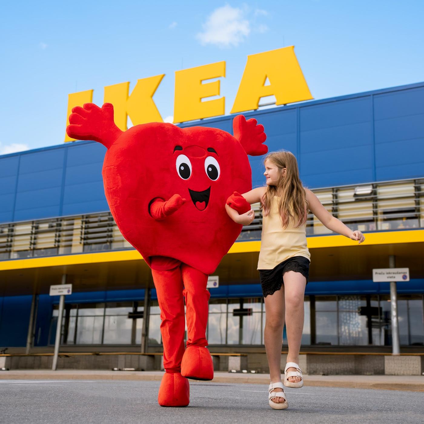A red Heart mascot and a girl in a yellow T-shirt and black shorts walk hand in hand, with an IKEA store in the background.