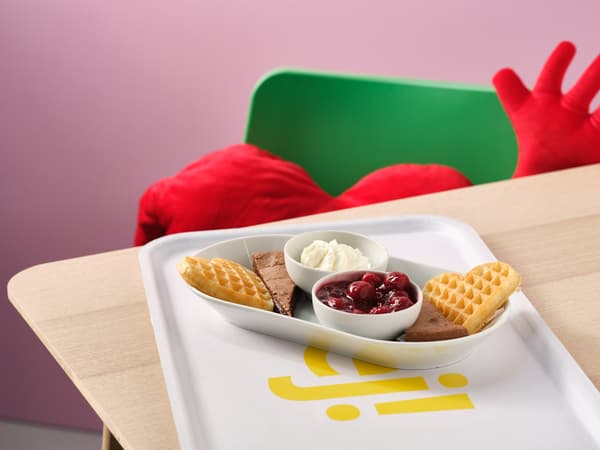 A red heart‑shaped cushion on a wooden table with an IKEA tray holding a special Valentine’s Day dessert in the Swedish restaurant, featuring heart‑shaped waffles, chocolate cake, compote and whipped cream.