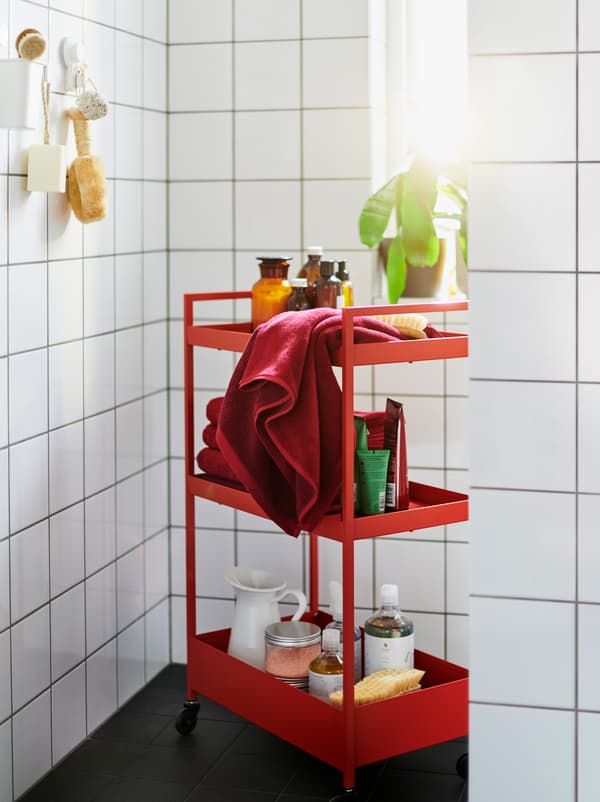 A red NISSAFORS trolley filled with accessories in a white-tiled bathroom also furnished with red and white ENHET modules.