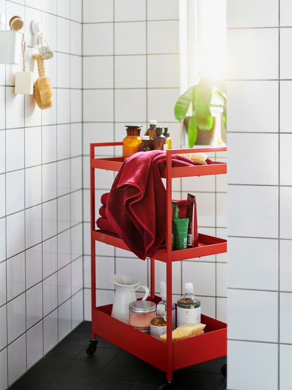 A red NISSAFORS trolley filled with accessories in a white-tiled bathroom also furnished with red and white ENHET modules.