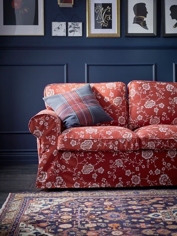 A red, patterned UPPLAND sofa with a plaid cushion in a living room with dark blue, panelled walls covered with framed art.