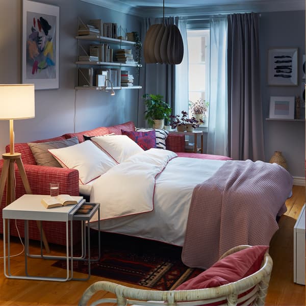A red sofa-bed made as a bed with white bed textiles, a white nest of tables and a wooden and white floor lamp.