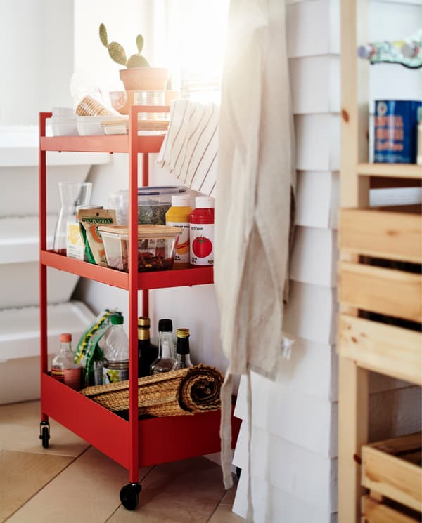 A red trolley on castors stands by a window. Food containers, sauces and bowls stand on the three trolley shelves.