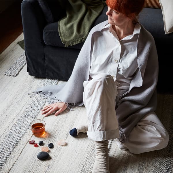 A redheaded woman sitting on a rug with a shawl over her shoulders, leaning on the sofa, beside a glass of tea and decorative stones.