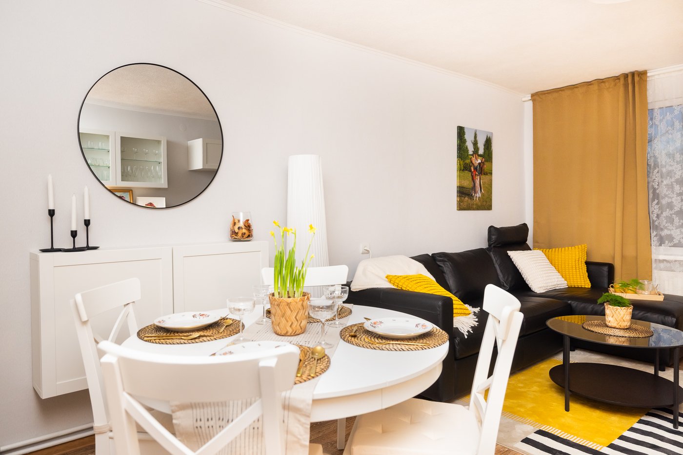 A renovated dining area with a round table, white chairs, and a large round wall mirror. The living area with a sofa and yellow accents is visible in the background.