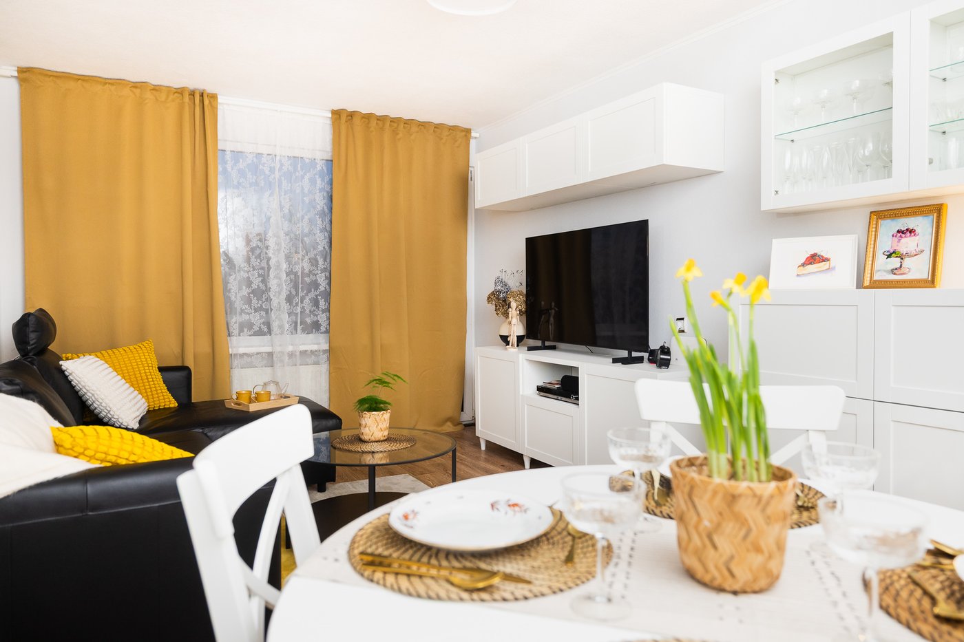 A renovated living room with light-coloured furniture, mustard-yellow curtains, and clearly defined zones – a dining area in the foreground and a seating area with a sofa and TV in the background. The space appears bright, tidy and cosy.