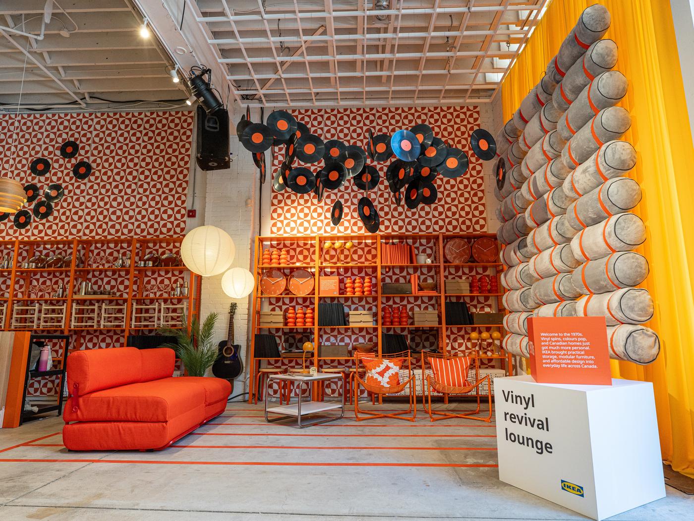 A retro-themed lounge with orange furniture, hanging vinyl records, patterned walls, shelves, and a sign reading “Vinyl revival lounge.”.