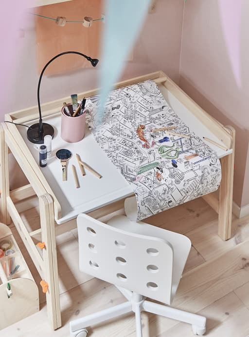 A roll of colouring paper on a child’s desk with a white chair against a pink wall.