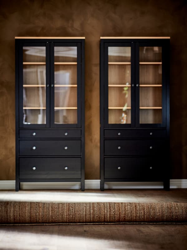 A rolled-up LOHALS rug lies on a wooden floor, a pair of empty glass-door cabinets stand against the brown wall behind.