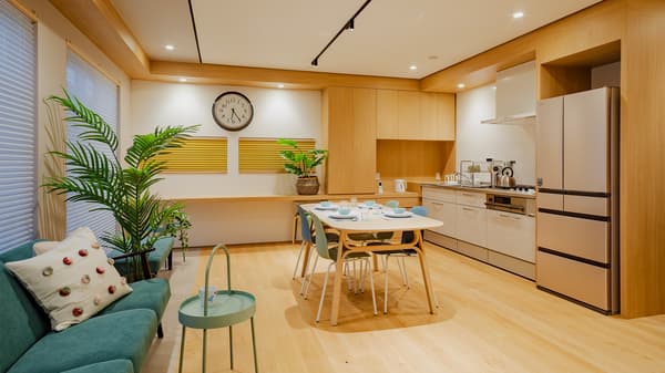 A room featuring a blue sofa in the foreground, a table and chairs in the background, and a built-in kitchen