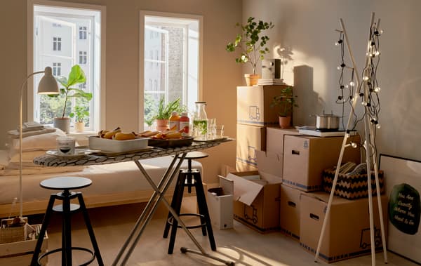 A room mid-moving in with stacked moving boxes, a small buffet served on an ironing board and a decorated EKRAR hat and coat stand.