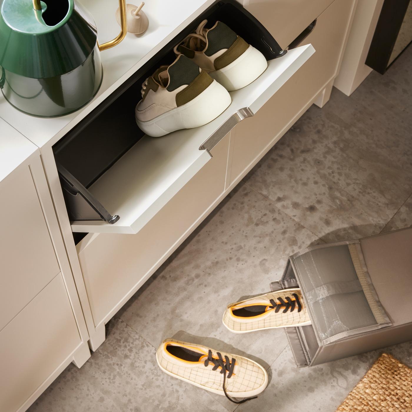 A room with a closed storage solution for shoes in white. There is an open drawer with slippers inside. There is a green watering can on top of the drawer.