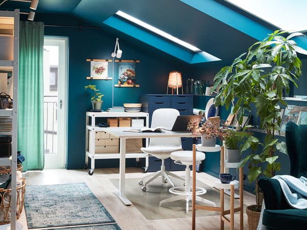 A room with a desk, a swivel chair and a sit/stand support in white, green plants on a plant stand and a green wing chair.