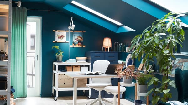 A room with a desk, a swivel chair and a sit/stand support in white, green plants on a plant stand.