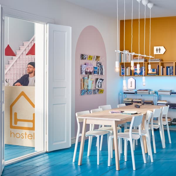 A room with a large wooden table and white chairs in the middle, and wall storage with magazines and books around it.