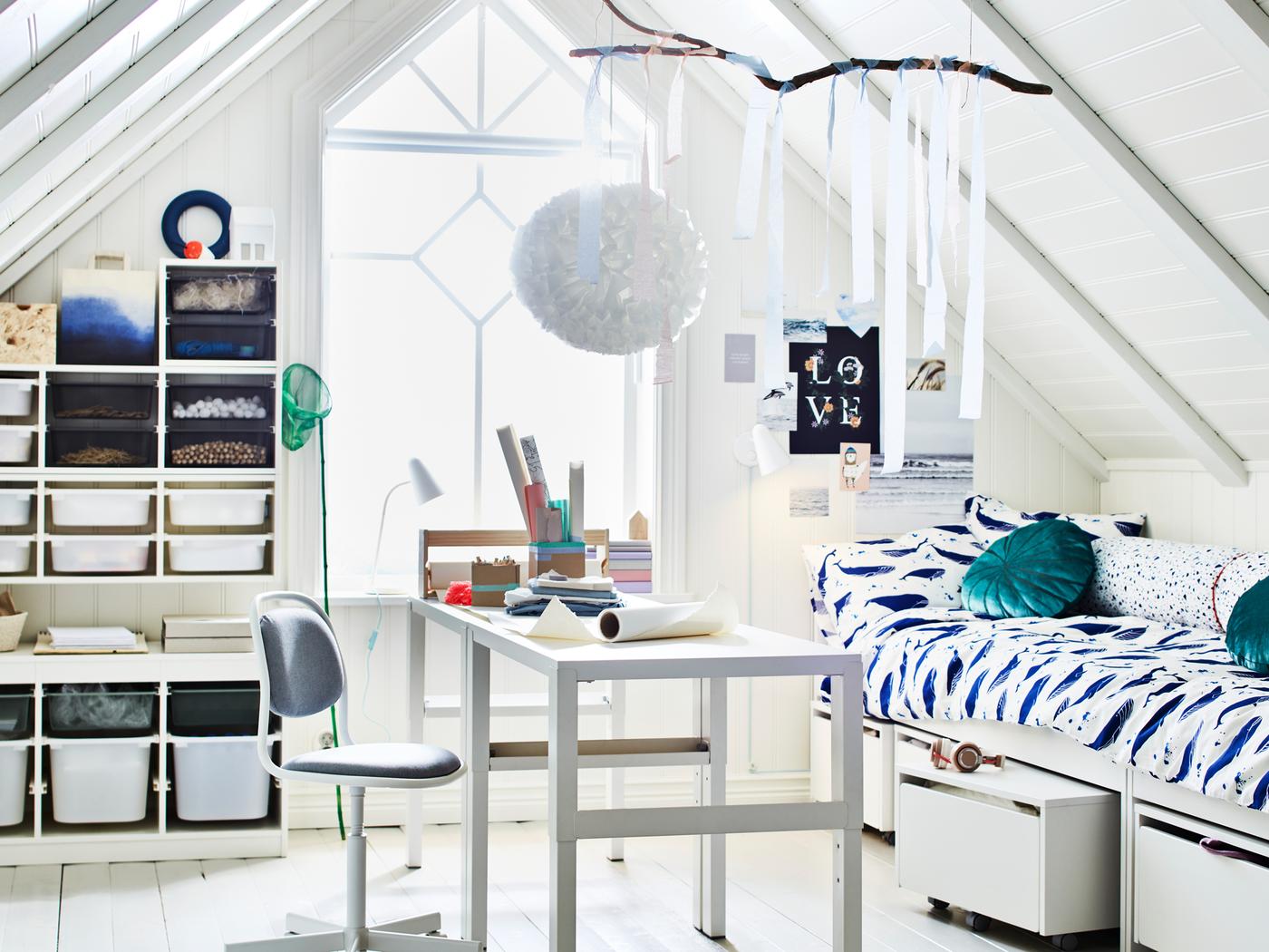 A room with a sloping ceiling and a large window. Two PÅHL desks stand in the middle near two beds and some TROFAST storage.