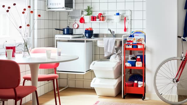 A room with a white SUNNERSTA mini-kitchen, a red trolley, TILLREDA kitchen appliances, a table with red chairs and a bike.