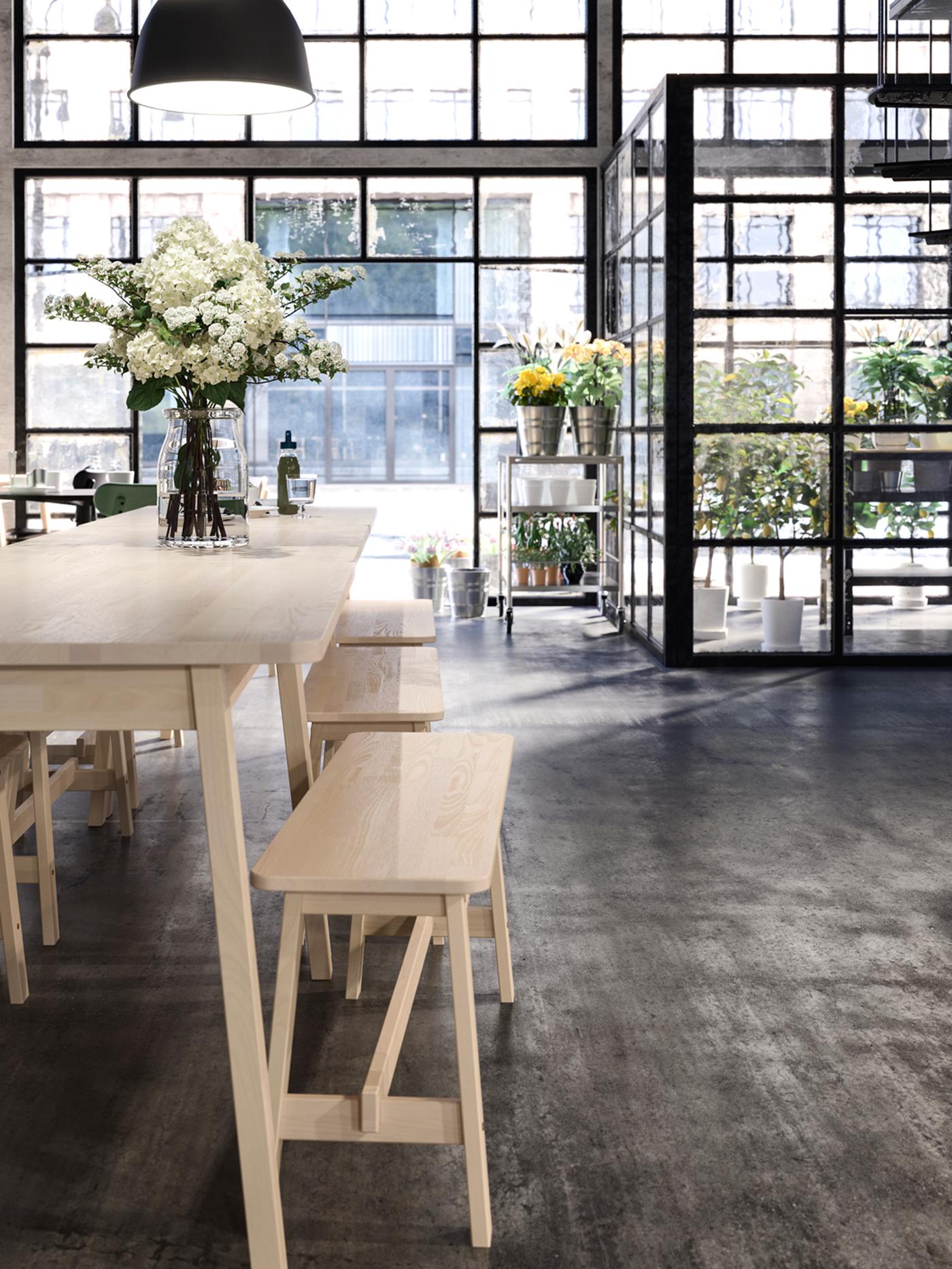 A room with a wooden table and chairs, a vase of flowers on the table, hanging lighting and windows in the background.