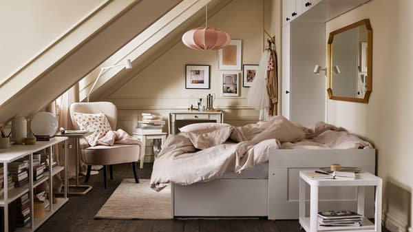 A room with an extended white daybed with light grey/beige bed linen, various storage, a beige armchair and pink pendant.