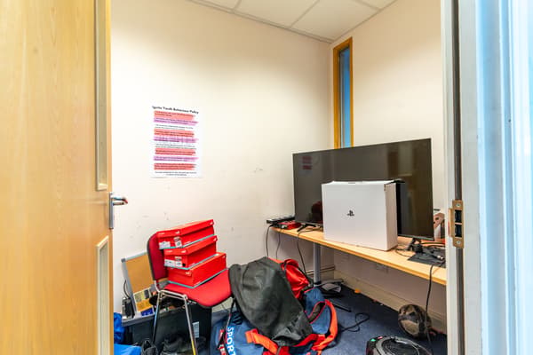 A room with beige walls, a wooden desk with a big screen on it, various red bags throughout.