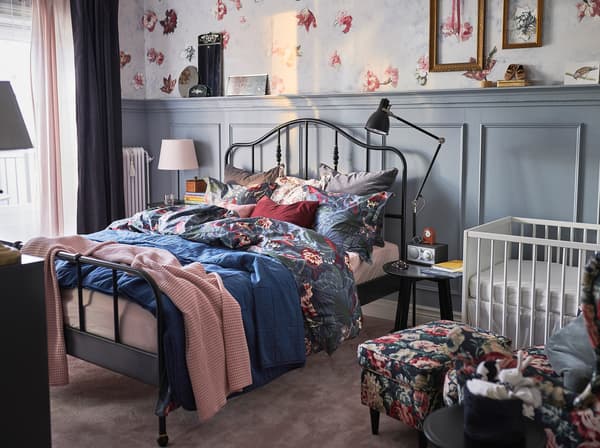 A room with floral patterns and pillows, a white crib and changing table, and a minimal black bedframe.