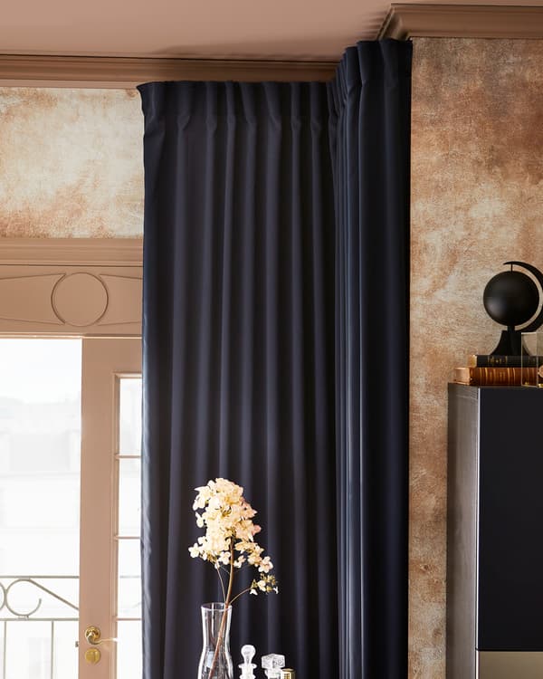 A room with MAJGULL block-out curtains in dark blue, drawn back to show the light, and a sprig of flowers in a glass vase.