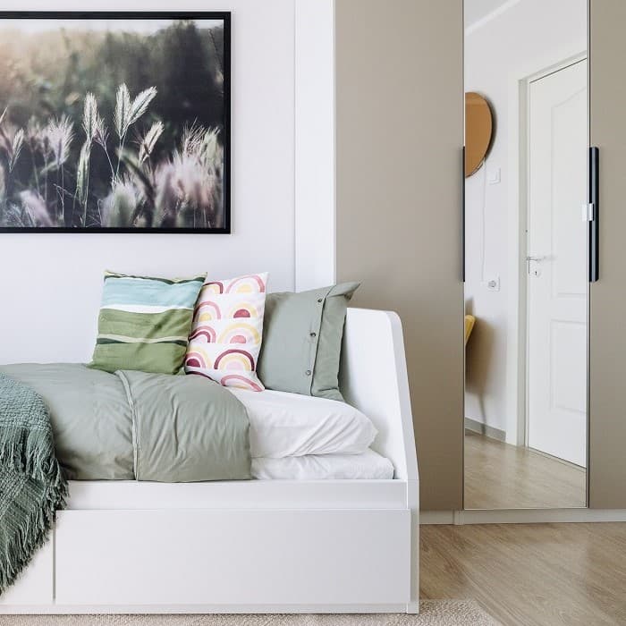 A room with white walls, a wardrobe with beige doors, a white bed with colorful pillows, and a floral picture on the wall.