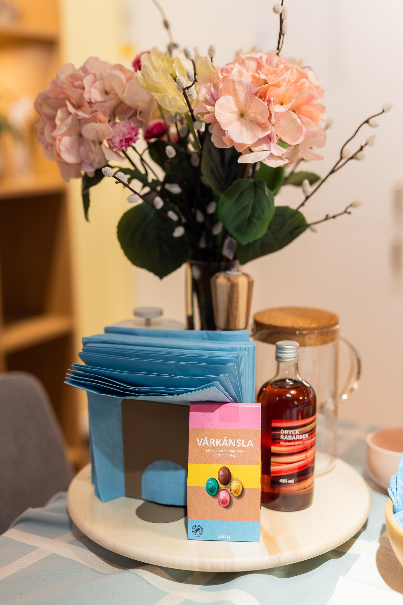 A rotating wooden tray holding napkins, a package of chocolate treats, and a bottle of syrup. A bouquet of pink flowers in the background.