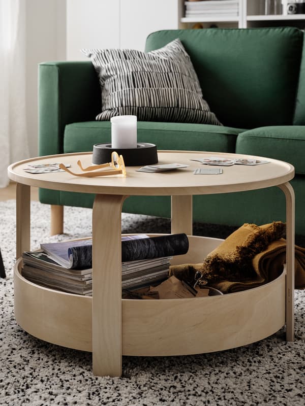 A round light wood BORGEBY table in front of a green sofa with playing cards, a block candle and a pair of spectacles on top.