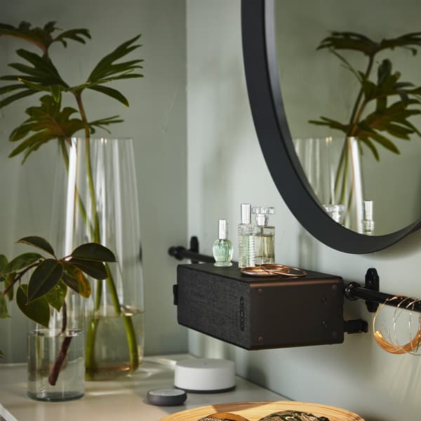 A round mirror, a black SYMFONISK WiFi speaker that hangs on a rail, plants in glass vases, some jewellery and perfumes.