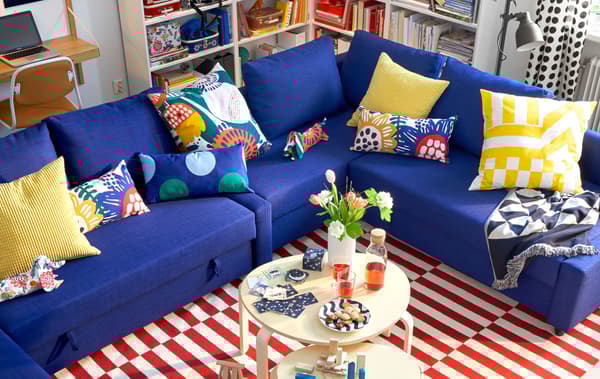 A royal blue FRIHETEN corner sofa with colourful cushions, two round coffee tables and a red and white striped rug.