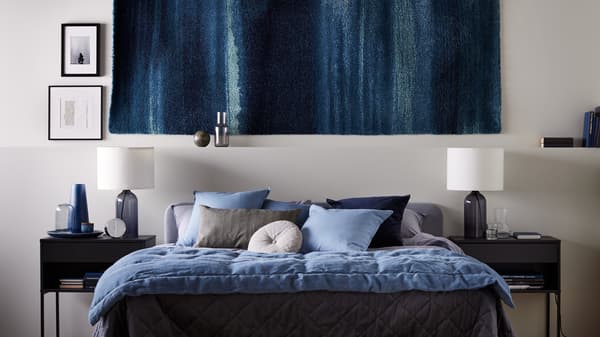 A rug on the wall, with VIKHAMMER nightstands either side of a light-gray SLATTUM full bed, with KOPPARBLAD bed linen.