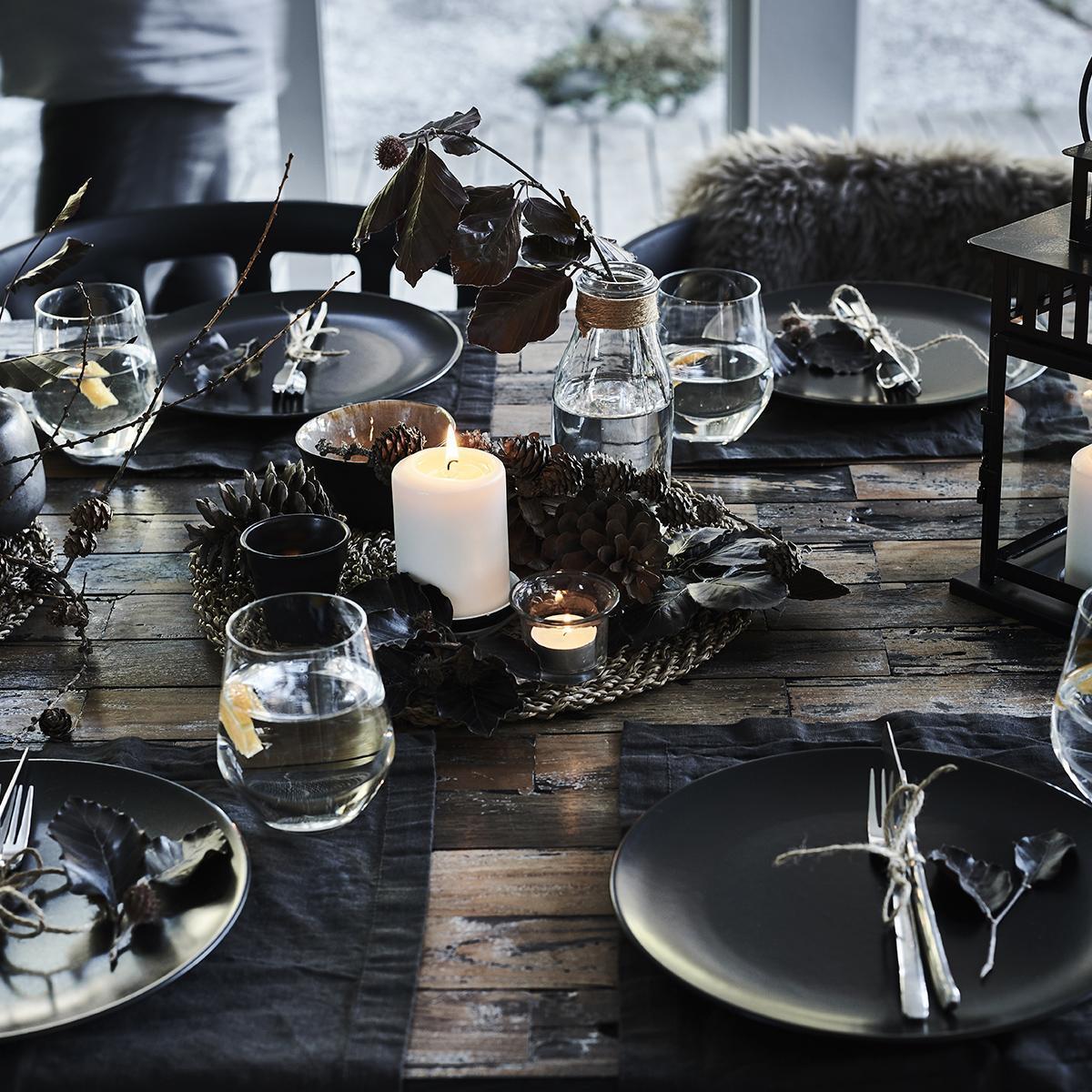 A rustic dining table with dark plates, glasses, pine cones and a burning candle, decorated with leaves and a cosy atmosphere.
