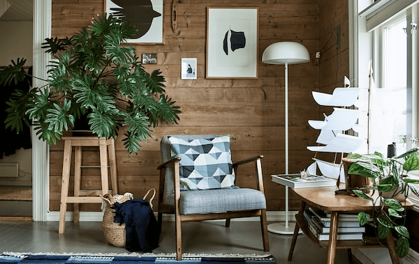 A rustic living room with a wood-framed armchair, white floor lamp and stool used as a plant stand for a big leafy plant.