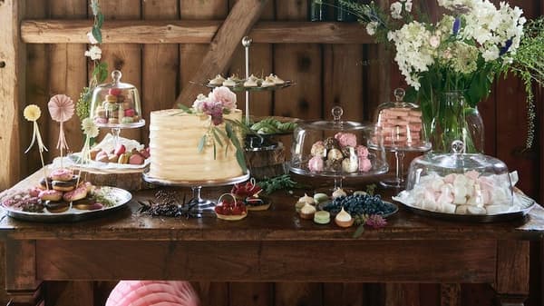 A rustic setting, a wooden table decorated with assorted cakes and treats and various fresh greenery.