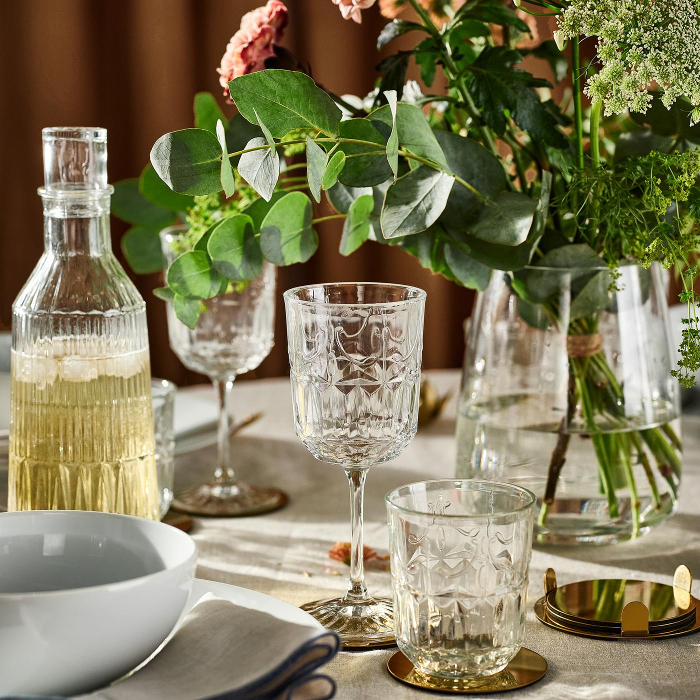 A SÄLLSKAPLIG glass carafe, glasses and wine glasses, plus flowers in a glass vase, on a table with a natural tablecloth.