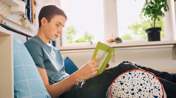 A school child sits reading on his bed, leaning back against a pillow with his legs supported by a MÖJLIGHET cushion.