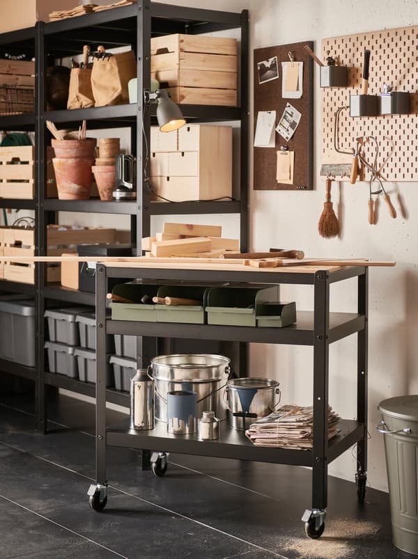 A section of a garage with a black BROR metal trolley with a wooden worktop and two shelves filled with boxes and paint cans.