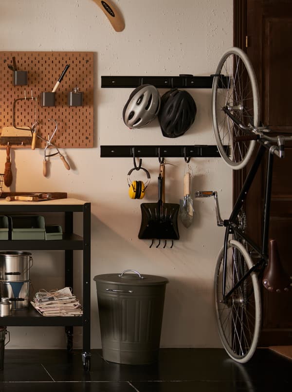A section of a garage with a grey KNODD metal bin with a lid and garden tools hanging on black metal wall hooks.
