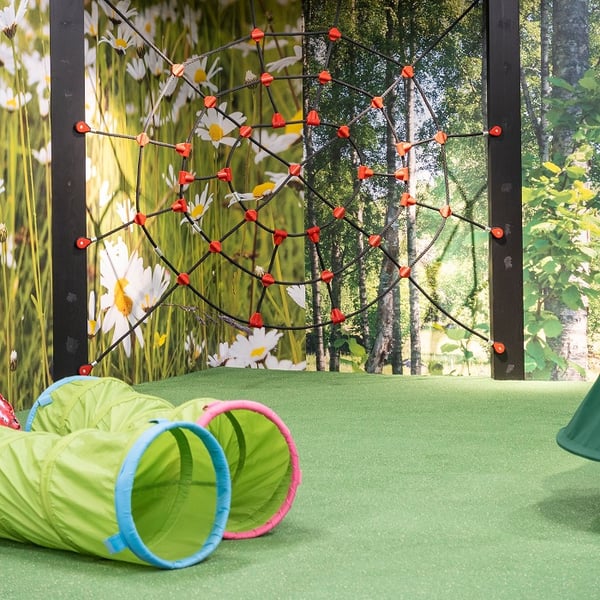 A section of an indoor children's play area featuring a spider web-style climbing net with red connectors mounted against a forest-themed wall mural, and two bright green crawl tunnels placed on soft green flooring.