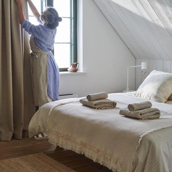 A serene bedroom with a cozy and rustic feel. A neatly made bed is covered with beige linens and topped with folded towels. A woman, dressed in a light blue shirt and apron, stands by the window adjusting the long beige curtains. The slanted ceiling features white wooden paneling, and a small white lamp sits on the bedside table. The room is softly lit by natural light coming through a green-framed window, with a small terracotta jug placed on the windowsill. A natural woven rug lies on the wooden floor, adding warmth to the space.