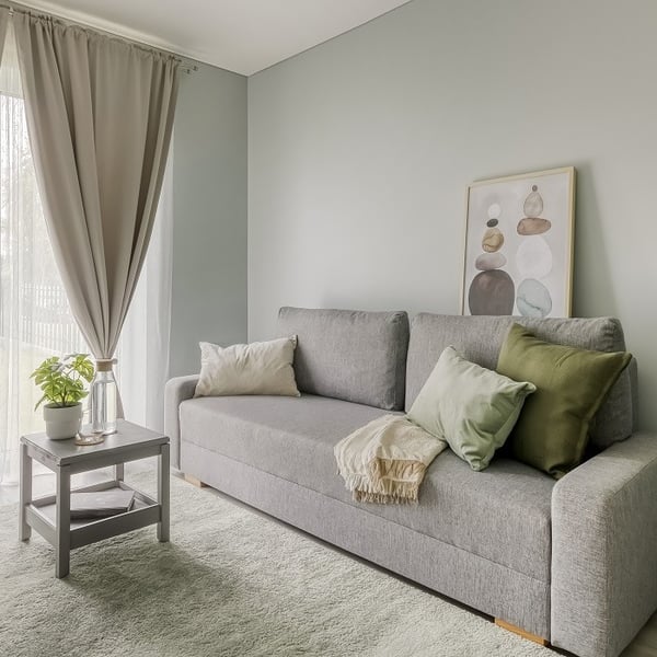A serene, light-filled sitting area featuring a modern gray sofa adorned with neutral and green throw pillows and a cream-colored blanket. A framed abstract art piece with stacked stones leans casually against the pastel green wall above the sofa. To the left, a small gray side table holds a potted plant and a glass carafe, adding a touch of freshness. Soft beige curtains and sheer white drapes frame the tall window, letting in natural light that highlights the cozy, green carpet underfoot. The space feels calm and inviting, ideal for relaxation or reading.