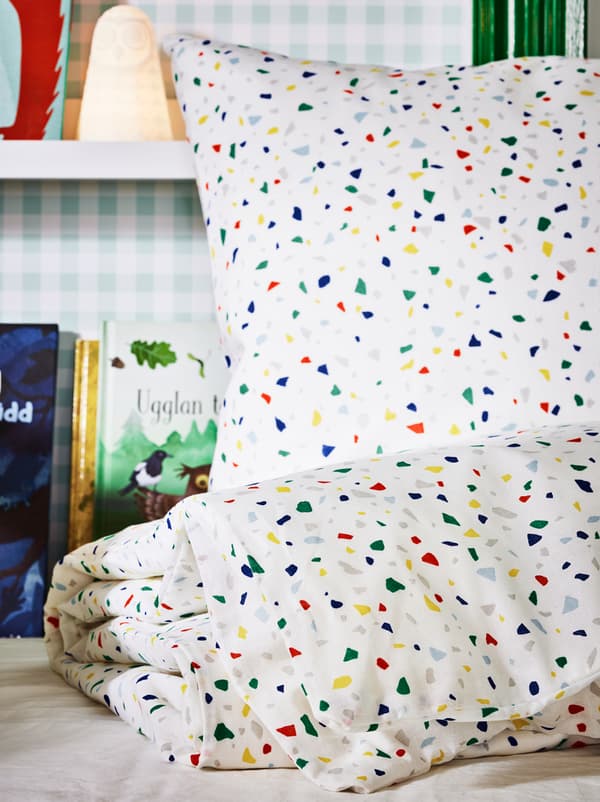 A set of MÖJLIGHET bed linen on an unmade bed, with a SOLBO LED lamp and books on picture ledges on a wall behind.