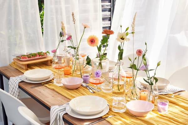 A set table in front of white curtains is set with plates, bowls and several vases with single flowers and grasses.