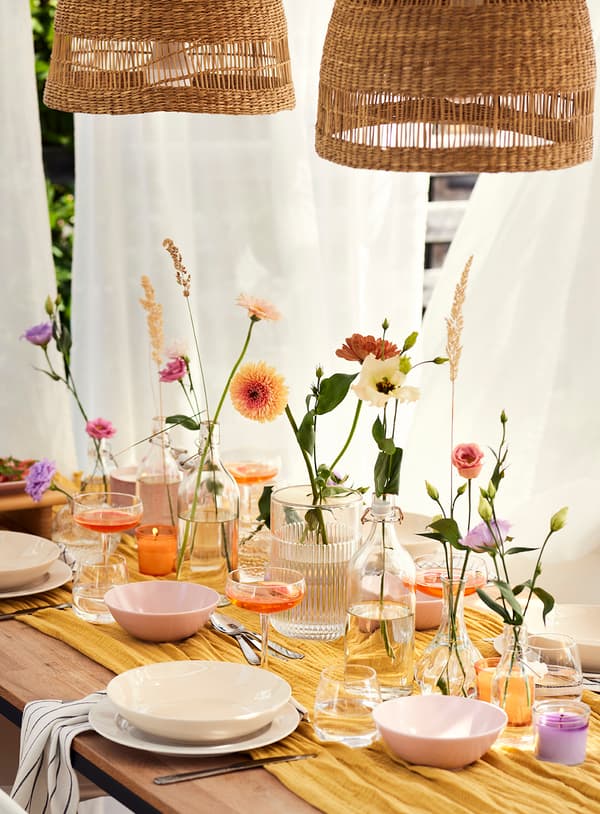 A set table with flower vases, glasses, and plates under two woven lampshades.