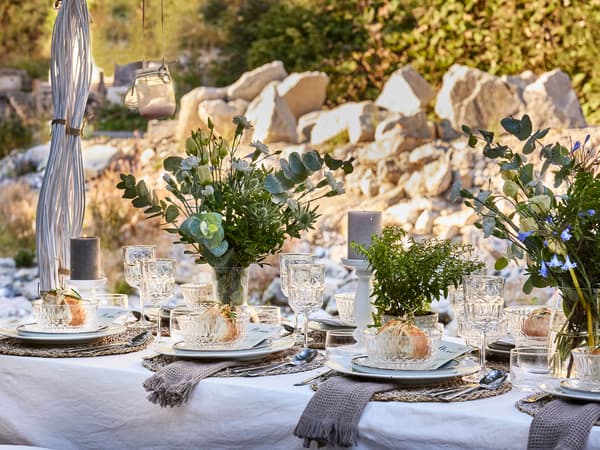A side view of a dining table set up with a white table cloth, grey round place mats, white dinnerware, various glasses and plants across. 