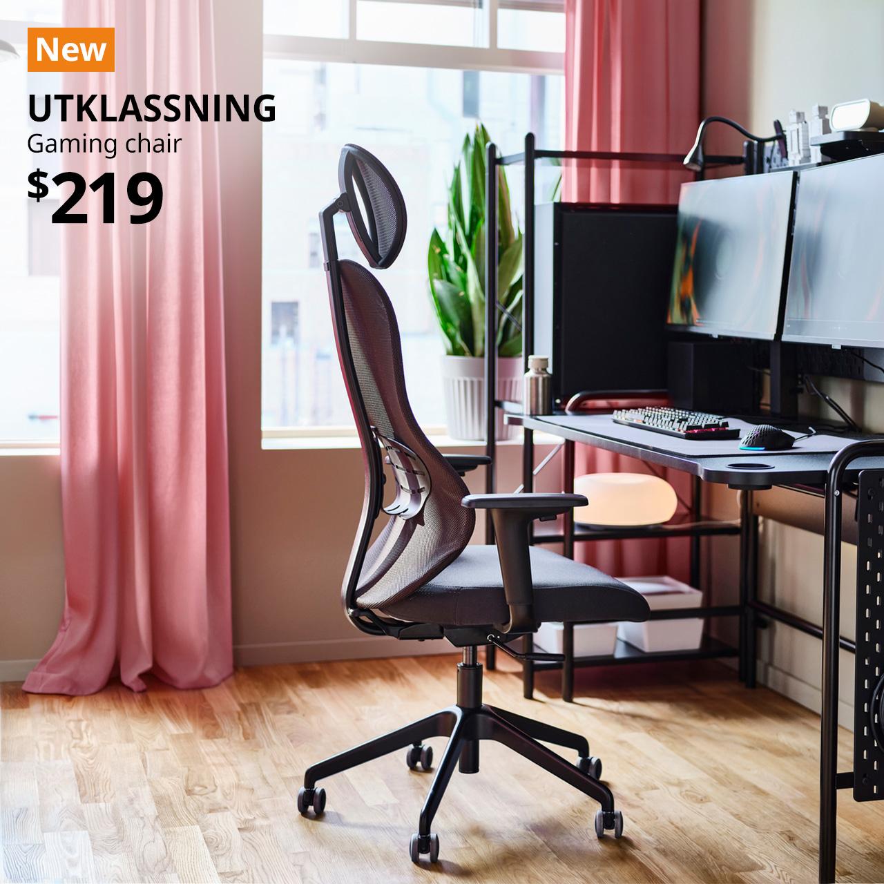 A side view of a Gräsnäs anthracite/black UTKLASSNING gaming chair in a home office and gaming setup corner in the living room. A orange tag with white text that reads 'New', followed by black text overlaying image that reads 'UTKLASSNING, Gaming chair, $219'.